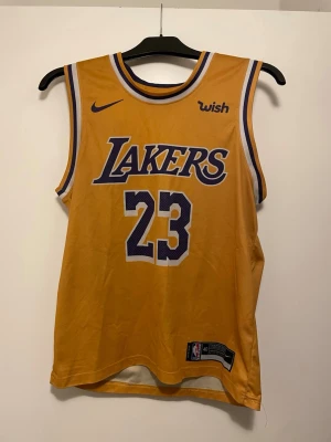 Gul Lakers basketlinne LeBron James - Snygg gul basketlinne från Nike med Lakers-logga och nummer 23, LeBron James. Linnet har lila och vita detaljer, är ärmlöst och tillverkat i ett lätt och ventilerande material. Perfekt för basketträning eller att bära till vardags för en sportig look.