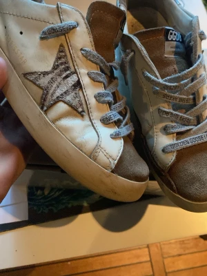 Golden Goose Superstar sneakers beige/brun - Golden Goose Superstar sneakers i beige och brunt med ikonisk stjärndetalj på sidan. Ovandel i läder och mocka, vita skosnören och högt skaft. Märkeslapp på plösen och vintageinspirerad design. Perfekta för dig som gillar streetstyle och unika detaljer.