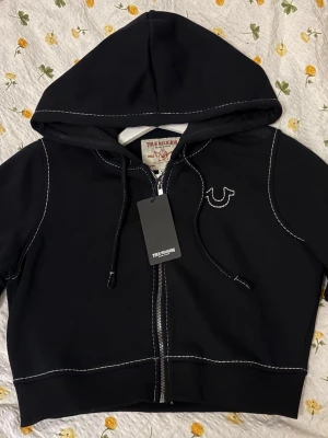 Svart hoodie från True Religion - Svart hoodie från True Religion med vit kontrastsöm, dragkedja och justerbar huva. Framtill finns logga broderad på bröstet och tryck bak på ryggen. Croppad modell i storlek L (passar mig som har S i hoodies) 