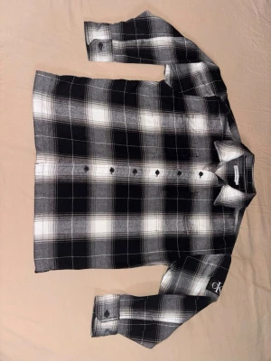 Rutig skjorta från Calvin Klein Jeans - Snygg svartvit rutig skjorta från Calvin Klein Jeans med klassisk krage och knappar framtill. Skjortan har lång ärm och logga broderad på ärmen. Perfekt för dig som gillar stilrena och tidlösa plagg. Ej Andvänd