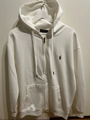 Vit hoodie från Polo Ralph Lauren - Snygg vit hoodie med dragkedja från Polo Ralph Lauren. Klassisk design med huva och dragsnören, två fickor framtill och den ikoniska lilla svarta loggan broderad på bröstet. Tillverkad i mjukt bomullsmaterial som är skönt att ha på sig.