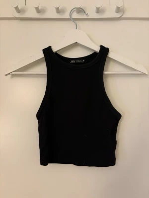 Svart croppad ribbad linnetopp Zara - Snygg svart croppad linnetopp från Zara med ribbad struktur. Toppen är ärmlös och har en tight passform med hög halsringning. Perfekt att matcha med höga jeans eller kjol för en trendig look. Storlek M men passar även XS