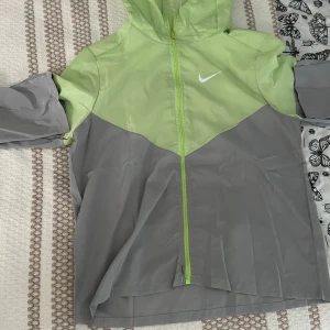 Nike Miler - Nike vindjacka med huva i fräsch limegrön och grå färgblock-design. Lätt och smidig i syntetmaterial. Har aldrig använt den eftersom den är för liten,  Storlek S, tveka inte på att höra av er 
