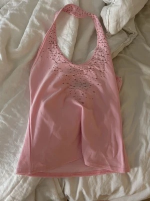 Rosa linnetopp med strassdetaljer - Säljer en ljusrosa linnetopp med halterneck och strassdetaljer framtill. Toppen är ärmlös och har en djup urringning. Perfekt för dig som gillar en feminin och glittrig stil.