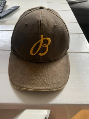 Brun keps från Breitling - Snygg brun keps från Breitling med gul broderad logga framtill och justerbart spänne bak. På baksidan står det 'Breitling 1884' i vitt och gult. Kepsen är i bomullstyg och har klassisk böjd skärm. Kepsen är lite solblekt från sommaren.