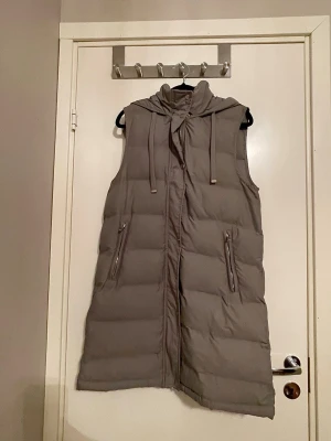 Lång grå dunväst med huva - ✨Snygg lång dunväst i grått med dragkedja framtill och två fickor med dragkedja. Västen har en stor huva med dragsko och quiltad design som ger en modern look. Perfekt att slänga över en hoodie eller tröja för extra värme ✨