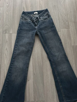 Blå bootcut jeans med broderad ficka - Säljer ett par blå bootcut jeans med snygg tvätt och dubbla knappar i midjan. Bakfickan har ett vitt broderat mönster som ger extra stil. Jeansen har klassiska fem fickor och kontrastsömmar. Perfekta för dig som gillar en avslappnad men trendig look.