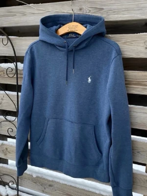 Blå hoodie från Polo Ralph Lauren - Snygg blå hoodie från Polo Ralph Lauren med klassisk huva och dragsko. Tröjan har en broderad vit logga på bröstet och en stor magficka framtill. Tillverkad i mjukt bomullsmaterial som känns skönt mot huden. Perfekt för en avslappnad och stilren look.