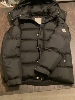 Svart Moncler vezere - Svart pufferjacka från Moncler med huva och ikonisk logga på ärmen. Jackan har quiltad design, dragkedja och knappar framtill samt ribbade muddar vid ärmslut. Insidan har ett coolt serietryck och innerficka med dragkedja. Perfekt för kalla dagar. Helt oanvänd köpte för ca 2 veckor sen och jackan passade inte mig så vill bli av med den snabbt Skriv för mer information/bilder .pris kan diskuteras vid snabb affär