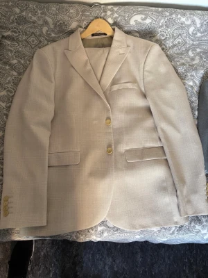 Beige kavaj med två knappar - Stilren beige kavaj från Lindbergh med två knappar framtill och klassisk krage. Kavajen har två fickor med lock, en bröstficka och fyra dekorativa knappar vid ärmsluten. Materialet är lätt strukturerat och ger en modern look.