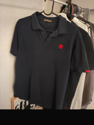 Svart pikétröja från Timberland - Klassisk svart pikétröja från Timberland med röd logga broderad på bröstet och liten röd detalj på ärmen. Tröjan har krage, knappar framtill och korta ärmar. Perfekt till jeans eller shorts för en avslappnad stil.