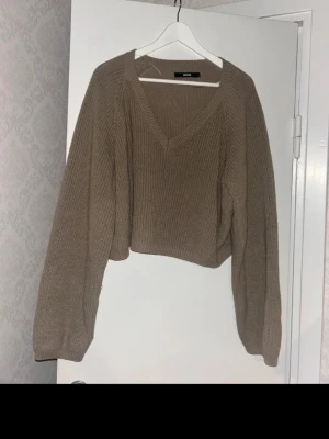 Beige cropped stickad tröja Bik Bok - Snygg beige stickad tröja från Bik Bok med croppad passform och djup V-ringning. Tröjan har långa, lite vidare ärmar och är ribbstickad för en trendig look. Perfekt att matcha med höga jeans eller kjol.