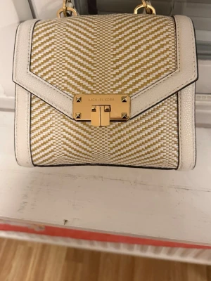 Beige och vit handväska Michael Kors - Äkta Snygg handväska från Michael Kors i beige och vitt med vävt mönster och vita kanter i läder. Väskan har guldfärgat lås framtill med logga och ett litet handtag upptill. Perfekt storlek för det viktigaste och en stilren accessoar till din outfit. 