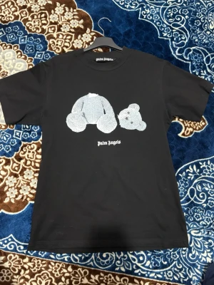 Svart Palm Angels t-shirt med björn - Svart t-shirt från Palm Angels med ikoniskt björntryck framtill och stor vit logga på ryggen. Klassisk passform och rund hals. Tillverkad i mjuk bomull som känns skön mot huden. Perfekt för dig som gillar streetwear och vill sticka ut.