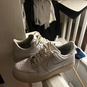 Nike Air Force 1 vita sneakers - Klassiska vita Nike Air Force 1 sneakers med låg profil och ikonisk swoosh på sidan. Skorna har perforerad tå för ventilation, tjock vit sula och ovandel i slätt läder. Perfekta för en clean och stilren look.