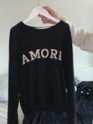 Svart stickad tröja Gina Tricot - Svart stickad tröja från Gina Tricot med vit text 'AMORE' framtill. Tröjan har rund halsringning och långa ärmar. Perfekt för dig som gillar enkel men snygg stil. Materialet känns mjukt och bekvämt.