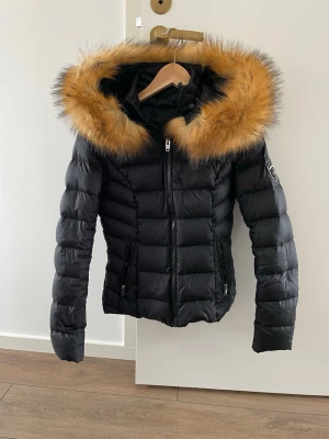 Svart dunjacka från ROCKANDBLUE med fuskpälsluva - Snygg svart dunjacka från ROCKANDBLUE med stor luva kantad av brun fuskpäls. Jackan har quiltad design, dragkedja framtill och två fickor med dragkedja. Märke syns på ärmen. Perfekt för kalla vinterdagar och ger en trendig look.