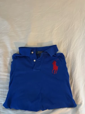 Blå långärmad polotopp från Polo Ralph Lauren - Klassisk blå långärmad polotopp från Polo Ralph Lauren med röd broderad logga på bröstet och röd siffra på ärmen. Topp med krage och knappar framtill, perfekt för en sportig och stilren look.