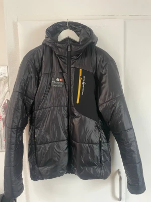 Svart dunjacka från sail racing  - Snygg svart dunjacka från sail racing med huva och gula detaljer på dragkedjorna. Jackan har en bröstficka med gul dragkedja och tryckt logga på bröstet samt en stor logga på huvan. Perfekt för vardagen. 