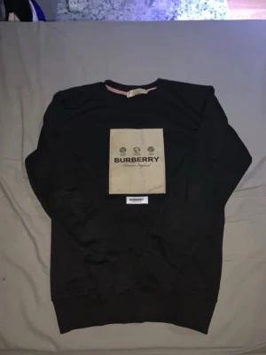 Svart sweatshirt från Burberry - Stilren svart sweatshirt från Burberry med stor beige patch och tydlig logga på bröstet. Tröjan har rund halsringning och ribbade muddar. Materialet ser ut att vara mjukt bomullstyg, perfekt för en trendig och avslappnad look.