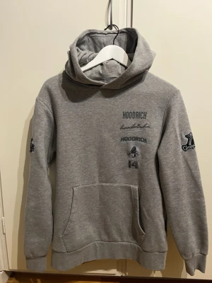 Grå hoodie från Hoodrich - Snygg grå hoodie från Hoodrich med tryck både fram och bak samt på ärmarna. Stor huva med broderad logga, känguruficka och ribbade muddar. Tillverkad i mjukt sweatshirtmaterial, perfekt för en chill och streetwear-inspirerad look.