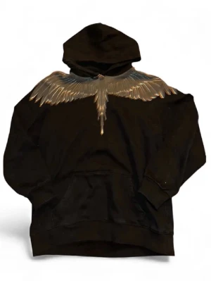 Svart hoodie Marcelo Burlon med vingar - Säljer en svart hoodie från Marcelo Burlon County of Milan med ikoniskt vingtryck i silver och guld över axlarna. Hoodien har huva och magficka, tillverkad i mjuk bomull för maximal komfort. Perfekt för dig som vill sticka ut med en unik design.