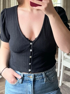 Svart ribbad topp  - Svart ribbad topp med v-ringning och korta puffärmar. Toppen har knappar framtill och är figurnära. Perfekt till vardags men även fest! Funkar med för storlek M