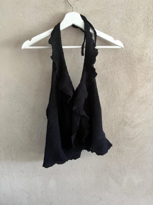 Svart halterneck topp med volang - Säljer en svart halterneck topp med volangkant och knyt i nacken. Toppen har en vågig struktur i tyget och djup urringning både fram och bak. Perfekt för dig som vill ha en unik och trendig look.