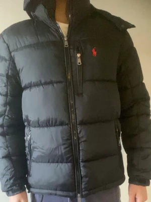 Svart dunjacka från Polo Ralph Lauren - Snygg svart dunjacka från Polo Ralph Lauren med röd broderad logga på bröstet. Jackan har hög krage, dragkedja framtill, två sidofickor med dragkedja och en bröstficka. Klassisk puffer-modell som håller dig varm under kalla dagar.