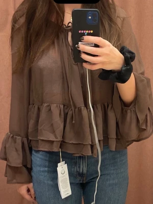 Brun volangblus med långa ärmar - Snygg brun blus i transparent material med volangdetaljer och knytning vid halsen. Blusen har långa, vida ärmar och en avslappnad passform som ger en trendig look. Perfekt att styla med jeans eller kjol för en cool outfit.