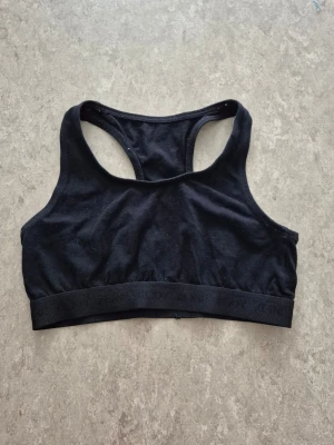 Sport-behå - En svart sport-bh från Kappahl med brottarrygg och bred resår nertill. Tillverkad i mjukt och stretchigt material som passar perfekt för träning. Diskret logga på resåren och klassisk, stilren design.