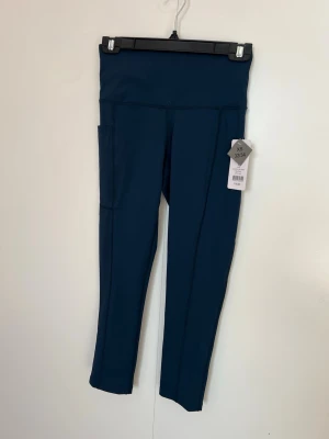 Nya,OPT träningsbyxor med hög midja i storlek XS(32-34) - Säljer ett par mörkblå leggings med hög midja och praktisk sidoficka på ena sida. De är tillverkade i ett stretchigt och mjukt material som sitter skönt på kroppen. Perfekta för träning eller chill dagar. Storlek XS (32/34).Djur och rökfritt hem 