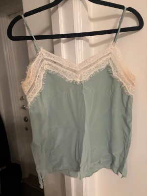 Mintgrön linnetopp med spetsdetaljer från Zara  - Supersöt Mintgrön linnetopp med tunna axelband och v-ringad spetskant i vitt. Toppen har dekorativa knappar bak och är perfekt för sommaren. Materialet känns lätt och svalt, och spetsen ger en romantisk touch.