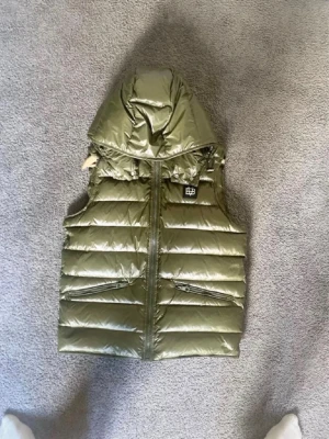 Bvaldi Olivgrön dunväst med huva - Snygg olivgrön dunväst med glansig finish och vadderad design. Västen har en stor huva, två dragkedjefickor framtill och en liten logga på bröstet. Perfekt för lager på lager och streetwear-vibe.