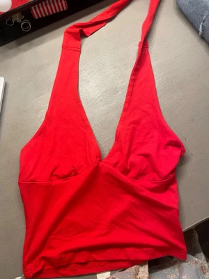 Röd halterneck linnetopp - Säljer en röd halterneck topp med djup v-ringning och breda band som knyts i nacken. Toppen är croppad och sydd i mjukt bomullstyg, perfekt för sommardagar eller festivaler. Enkel men snygg design som verkligen sticker ut.