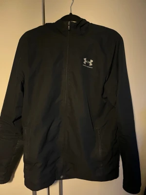 Svart vindjacka från Under Armour - Snygg svart vindjacka från Under Armour med dragkedja framtill och huva. Jackan har två sidofickor med dragkedja och ett meshfoder på insidan för extra komfort. Perfekt för blåsiga dagar och sportiga outfits. Logotypen syns tydligt på bröstet.