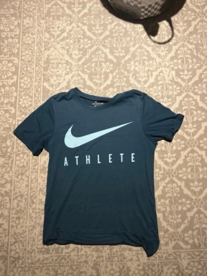 Blå Nike Dri-Fit T-shirt Athlete - Sportig blå T-shirt från Nike med stort vitt Swoosh-tryck och texten ATHLETE på bröstet. Tillverkad i Dri-Fit material som andas och håller dig torr under träning. Klassisk passform och rund hals.