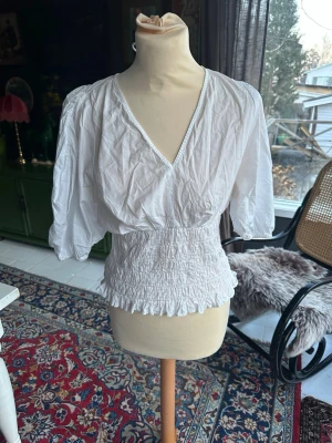 Vit smockad blus med v-ringning - Snygg vit blus med djup v-ringning fram och bak samt smockad midja som ger en feminin siluett. Blusen har trekvartsärm och knytband i nacken. Tillverkad i ett lätt och luftigt tyg, perfekt för en trendig och avslappnad look.