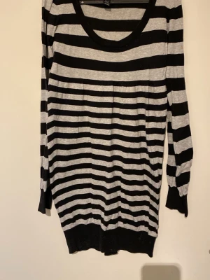 Randig stickad tröja från H&M - Stickad tröja från H&M med breda svarta och gråa ränder. Tröjan har rund halsringning, långa ärmar och ribbade muddar vid ärmslut och nederkant. Perfekt för en avslappnad och trendig look.