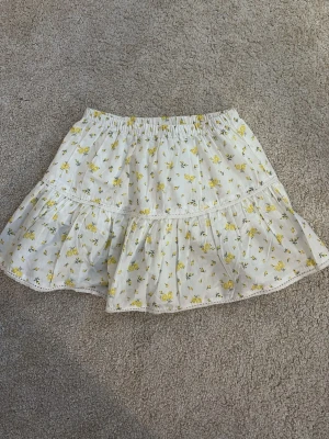 Vit gul blommig kortkjol med volang - Supersöt vit kortkjol med små gula blommor och volangkant. Kjolen har resår i midjan och fina spetsdetaljer längs sömmarna. Perfekt för varma dagar och ger en fräsch och somrig vibe. Mycket bra skick och är endast använd ca 1-2 gånger. 