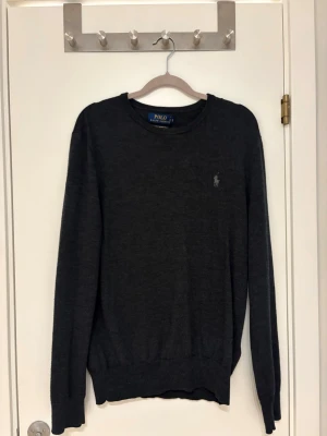 Mörkgrå tröja från Polo Ralph Lauren - Mörkgrå långärmad tröja från Polo Ralph Lauren med rund halsringning och diskret broderad logga på bröstet. Tröjan är tunnstickad och har ribbade muddar vid ärmslut och nederkant. Perfekt för en stilren och klassisk look. 100 procent merinoull| storlek m| skick 9/10