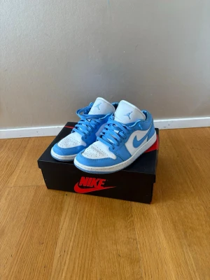 Nike Air Jordan 1 Low  blå/vit - Snygga Nike Air Jordan 1 Low i klassisk UNC-färg. Skorna har en fräsch kombination av ljusblått och vitt läder, med blå snörning och den ikoniska swoosh-loggan på sidan. Perfekta för dig som vill sticka ut med en stilren och sportig sneaker.