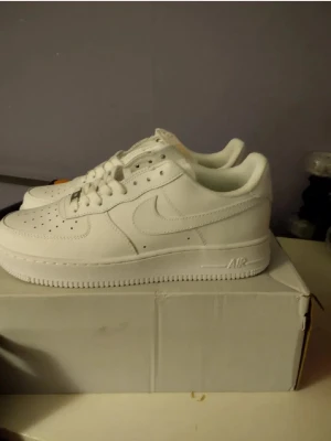 Nike Air Force 1 vita sneakers - Klassiska helvita Nike Air Force 1 sneakers i läder med perforerad tå, tjock sula och ikonisk Swoosh-logga på sidan. Skorna har lågt skaft och vita skosnören. Perfekta för en clean och stilren look.