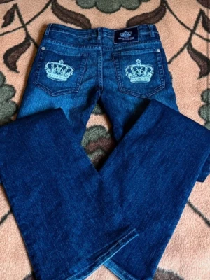 Mörkblå  Victoria Beckham jeans  - Snygga mörkblå jeans från Victoria Beckham  med utsvängda ben och broderade kronor på bakfickorna. Klassisk femficksmodell i denim med låg midja och detaljer i ljusblått broderi. Perfekta för dig som gillar statement-plagg och streetstyle.