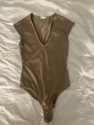 Beige body med djup v-ringning - Snygg beige body med djup v-ringning och korta ärmar. Tillverkad i ett stretchigt och mjukt material som sitter tight mot kroppen. Perfekt att styla med jeans eller kjol för en trendig look.