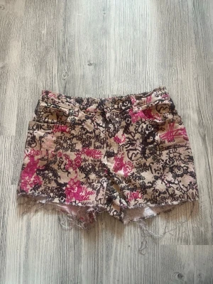 Beige shorts med graffiti och rosor - Unika shorts i beige med svart och rosa graffiti- och rosmönster. Råa kanter nertill ger en avslappnad vibe. Klassisk femficksmodell med bälteshällor och knappstängning. Perfekta för dig som vill sticka ut i sommar.