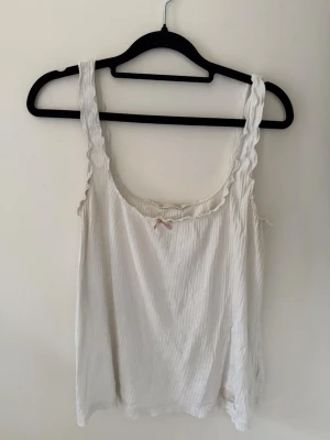 Vit ribbad linnetopp från Odd Molly  - Söt vit linnetopp med ribbad struktur och volangkant runt halsringningen. Toppen har en liten rosett framtill och breda axelband. Perfekt för varma dagar och enkel att styla med jeans eller kjol.
