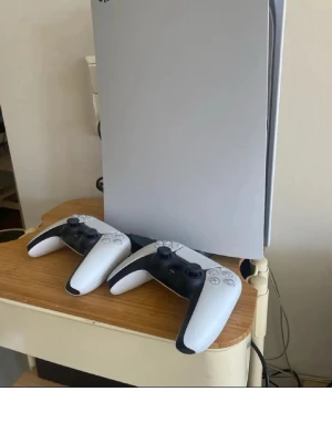 PlayStation 5 med två DualSense-kontroller - PlayStation 5 konsol i vit och svart design från Sony. Medföljer två matchande DualSense-kontroller i samma färg. Konsolen har en modern, stilren form det är ps5 digital edition slim