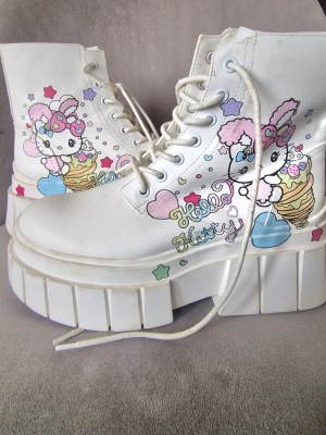 Hello Kitty vita platåstövletter - Unika vita platåstövletter med Hello Kitty-motiv i pastellfärger. Skorna har höga, chunky sulor och snörning framtill. Dekorerade med färgglada stjärnor, hjärtan och glassar. Tillverkade i konstläder och perfekta för dig som älskar kawaii-stil.