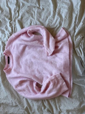 Ljusrosa stickad tröja - Mysig ljusrosa stickad tröja med rund halsringning och ribbade muddar vid ärmslut och nederkant. Tröjan har en avslappnad passform och är perfekt för att hålla värmen under kyliga dagar. Materialet känns mjukt och behagligt mot huden.